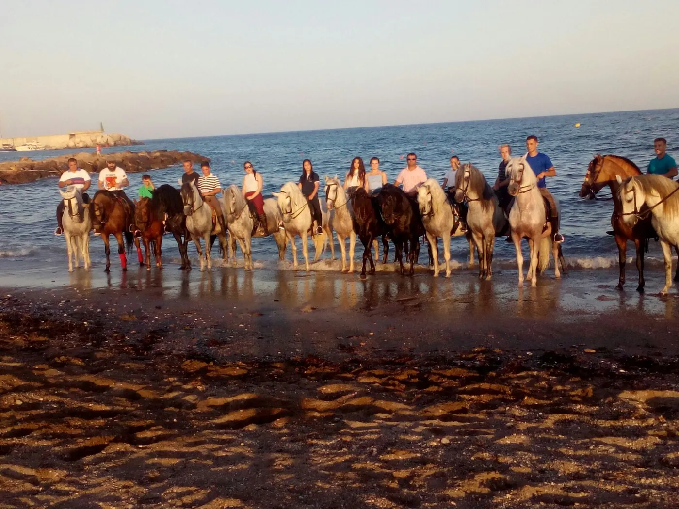 Grupo del centro hípico montando dentro del agua en la playa al atardecer